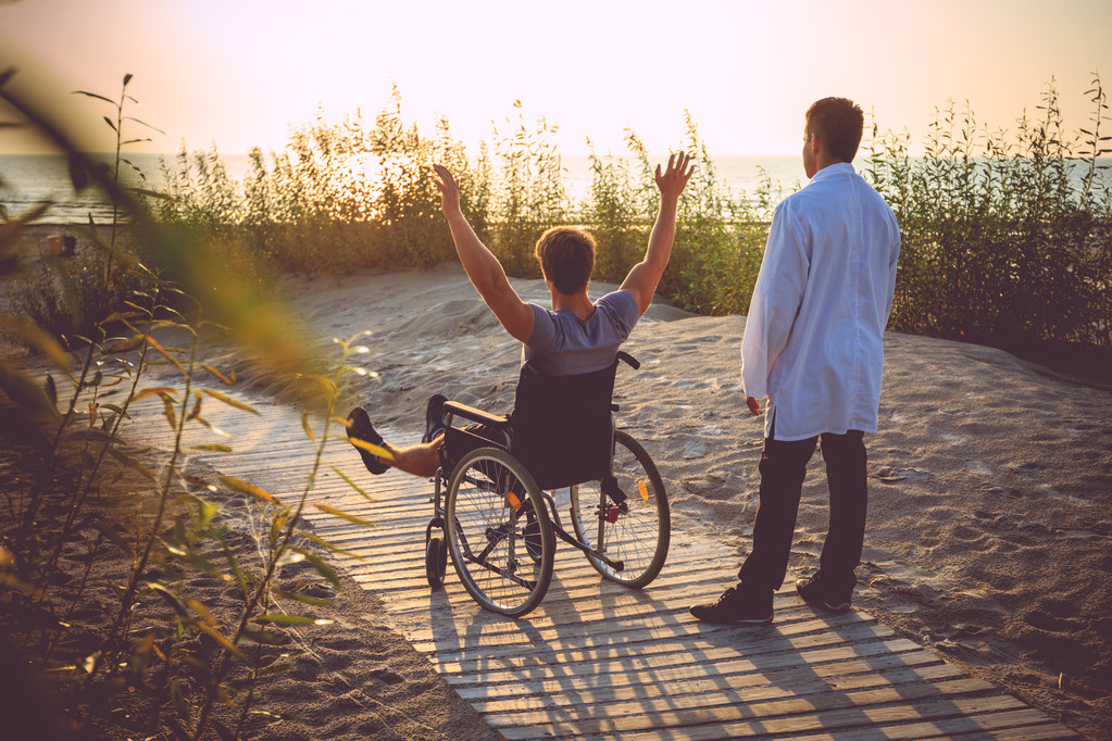 A man on wheelchair and his nurse enjoying sunrise on a beach.
